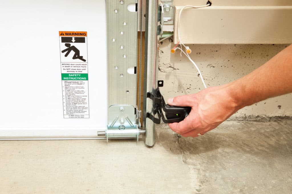 A hand is adjusting a garage door safety reversing sensor, with an illuminated green LED to confirm the sensor is lined up with the opposite side sensor and operational. These sensors, required in all new installations, reverse the door when an infrared beam is broken by a child, animal or object. A warning and safety instructions sticker is to the left. There is some paint overspray visible on the sensor and track.